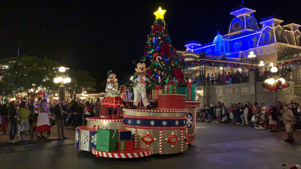 a group of people in clothing on a parade float with a christmas tree and a lit up building