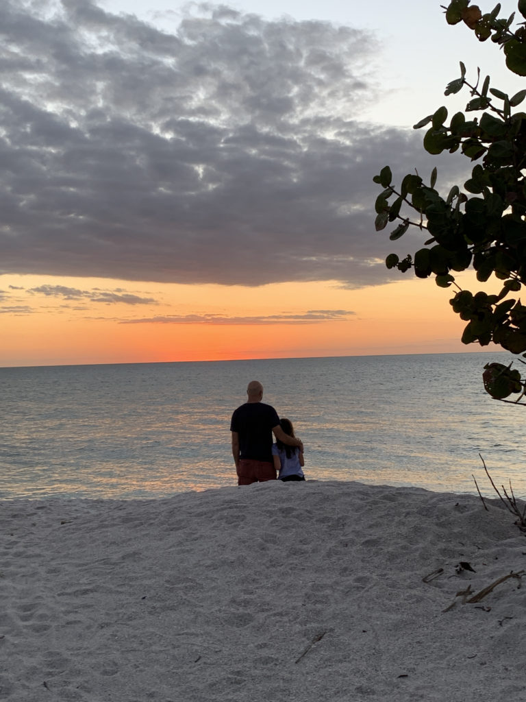 a man and woman standing on a beach looking at the sunset