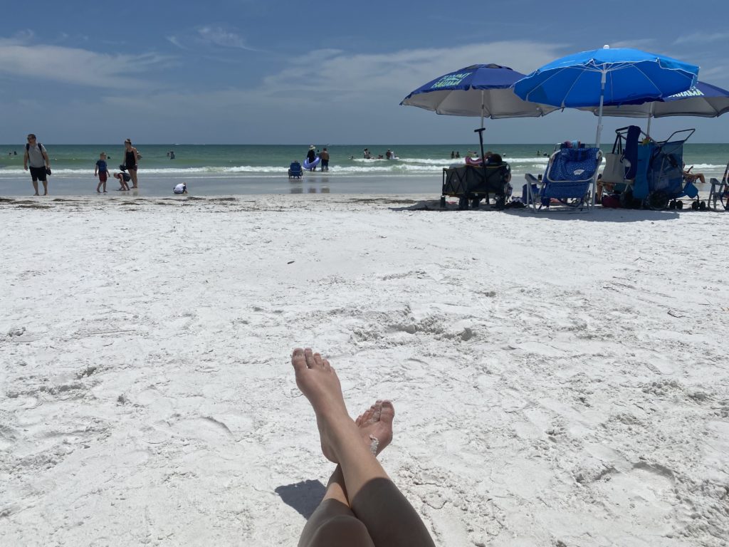 a person's legs on a beach with umbrellas and people in the background