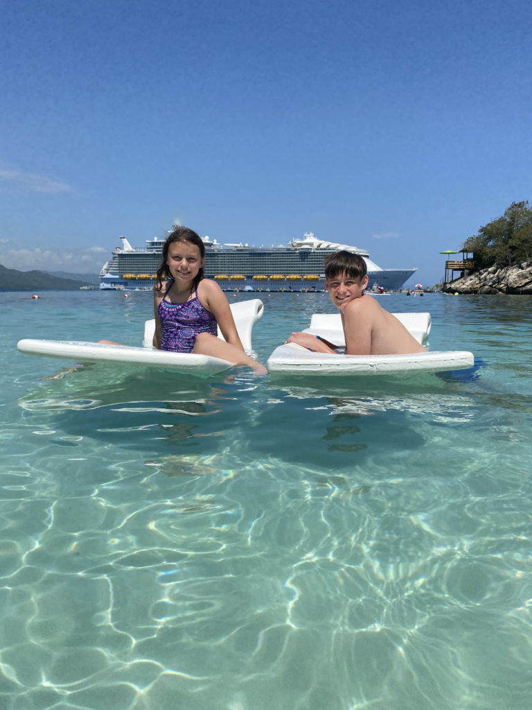 a boy and girl on lounge chairs in water with a cruise ship in the background