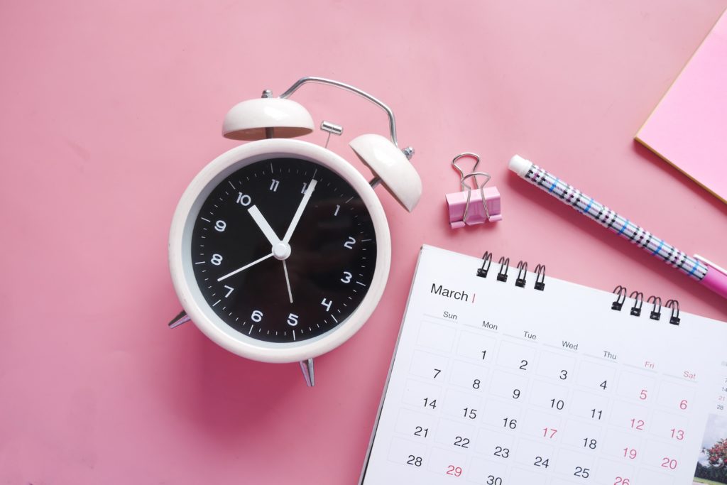 a white alarm clock next to a calendar and a pen