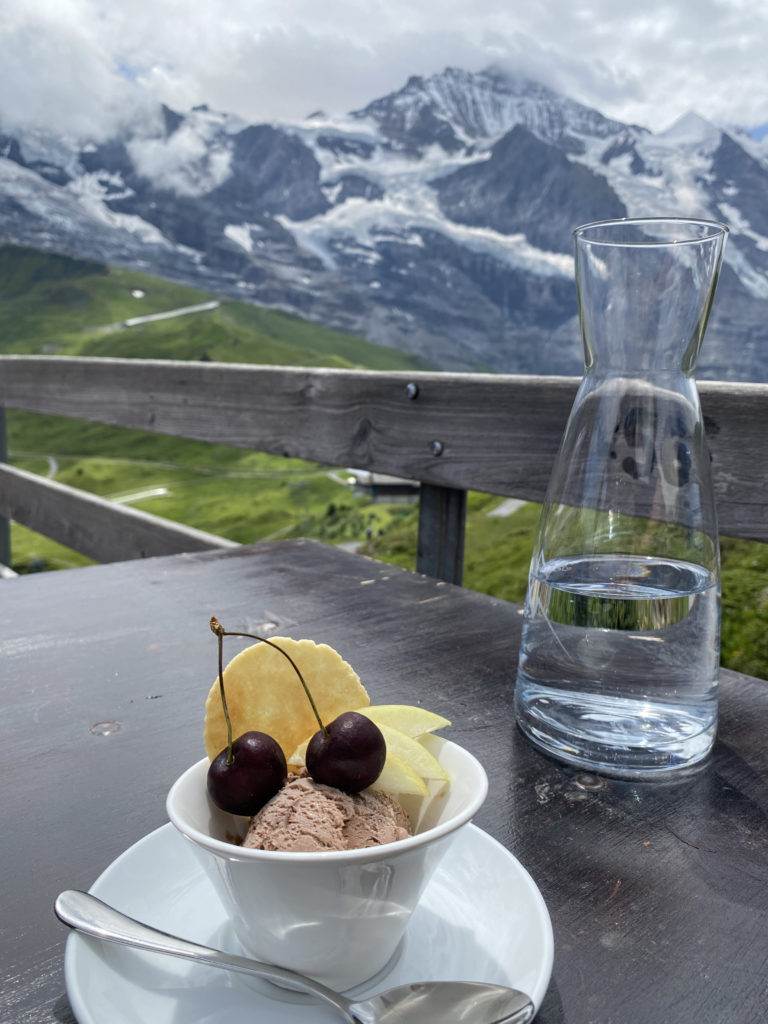a bowl of ice cream and fruit on a table with a glass of water and mountains in the background
