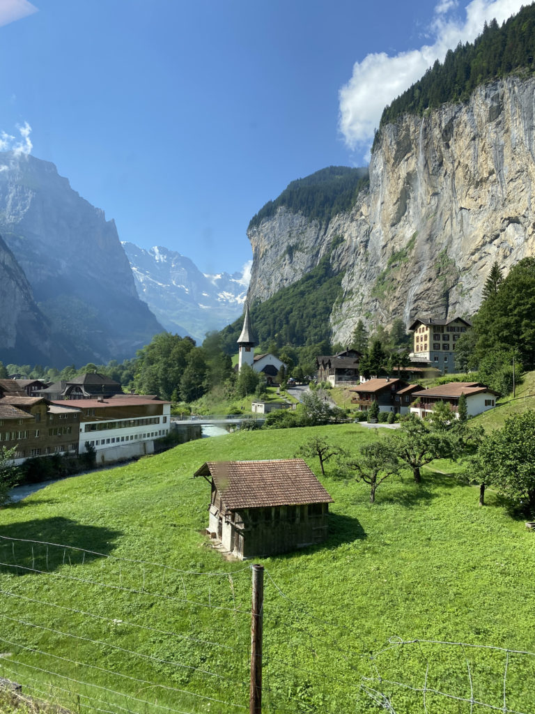 a green field with buildings and a river in front of a cliff