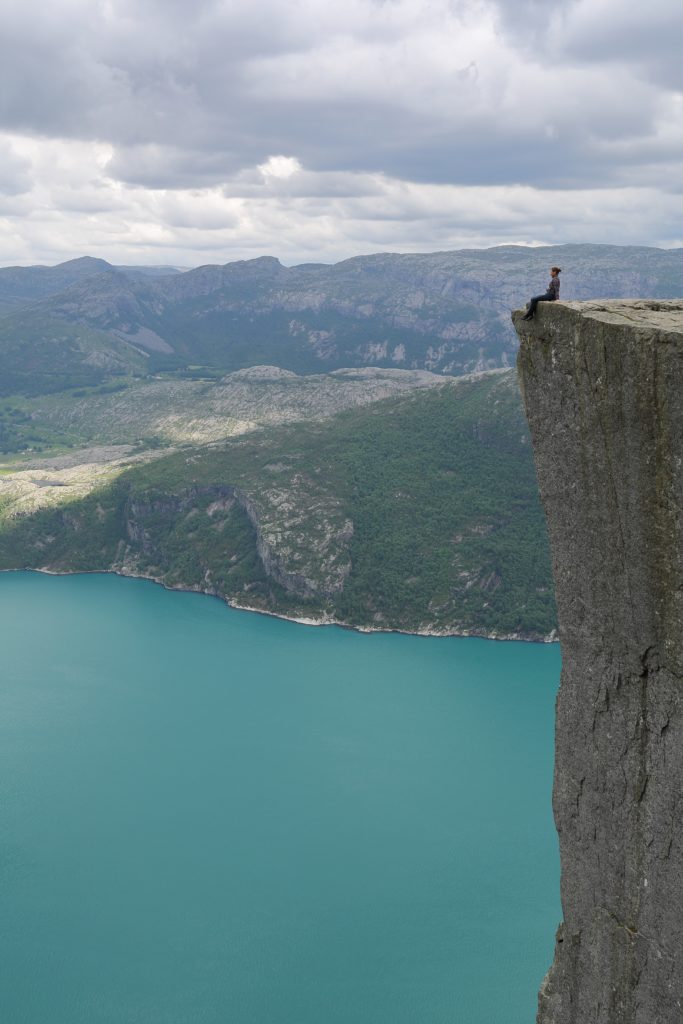a person sitting on a cliff edge overlooking a body of water with Preikestolen in the background