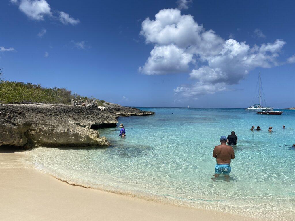 A scenic beach with clear turquoise water and a sandy shore. Several people are enjoying the water, and a sailboat is anchored in the distance. The sky is blue with a few fluffy clouds, and rocky cliffs are visible on the left side of the image.