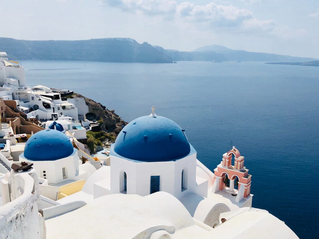 a white and blue buildings with blue domes overlooking the water with Santorini in the background