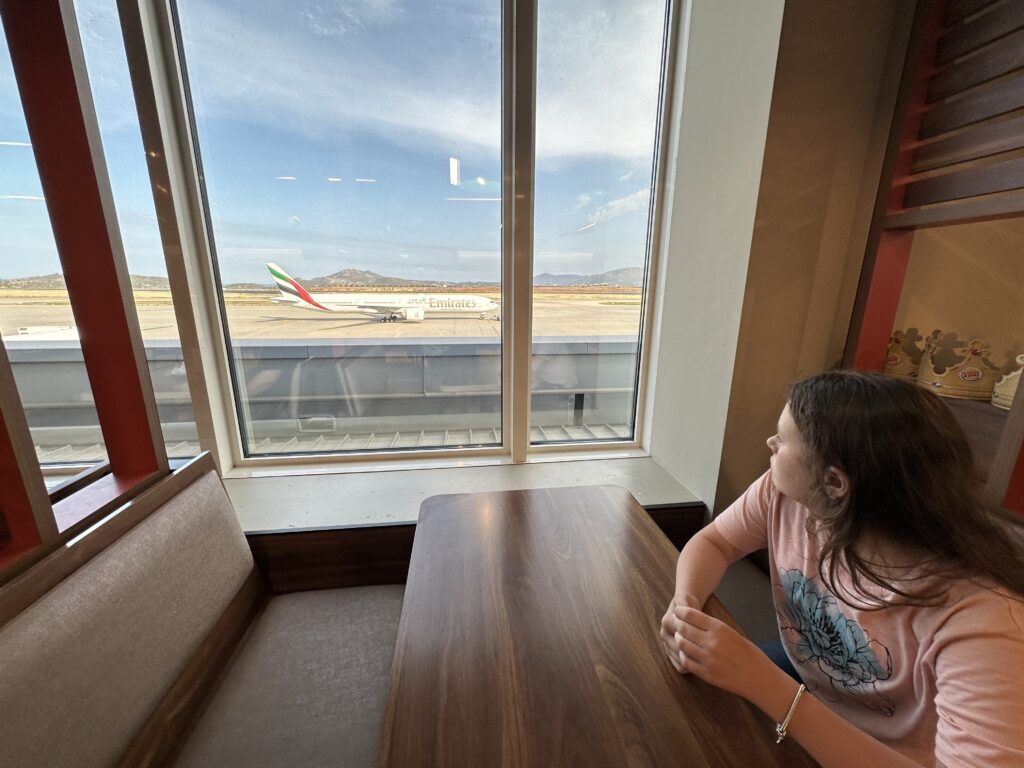 a girl sitting at a table looking out a window at an airplane