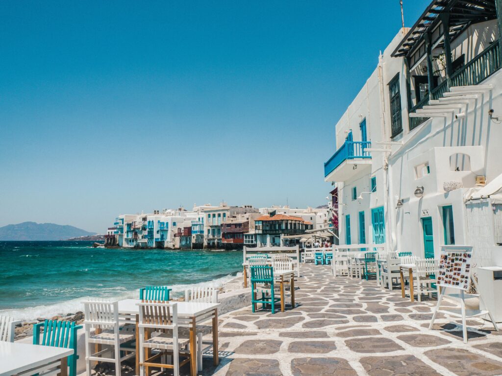 a white building with blue windows and tables on a stone walkway by the water
