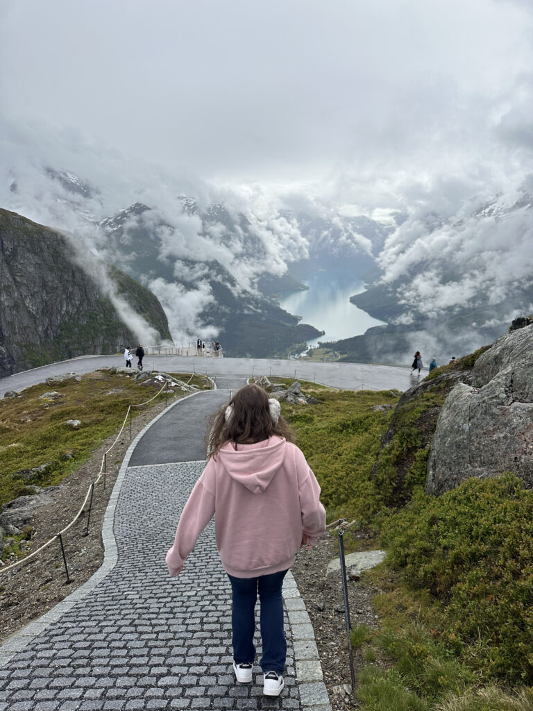 a girl walking down a path on a mountain with a river and clouds