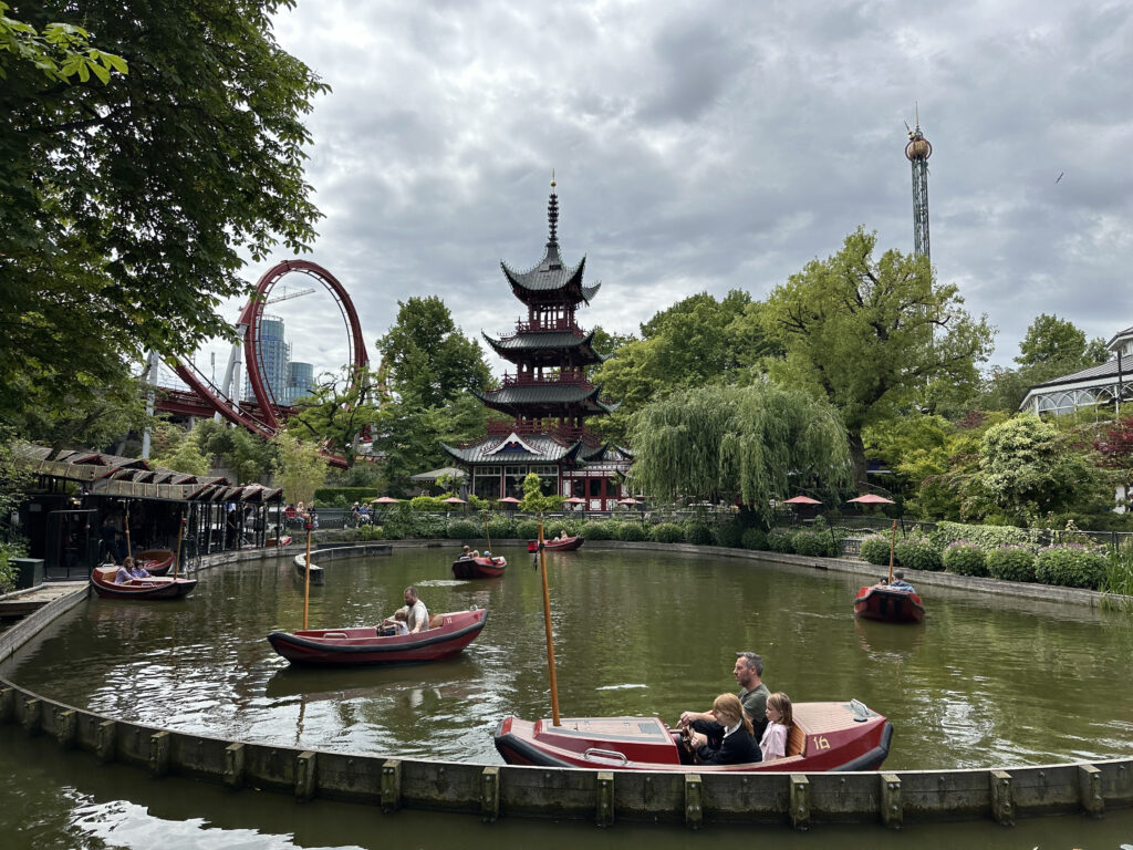 a group of people in a boat on a pond with a red roller coaster and trees with Tivoli Gardens in the background