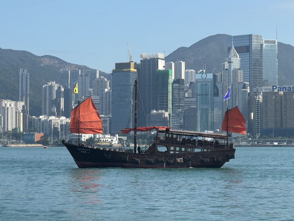 a boat with red sails in water with city in the background with Victoria Harbour in the background