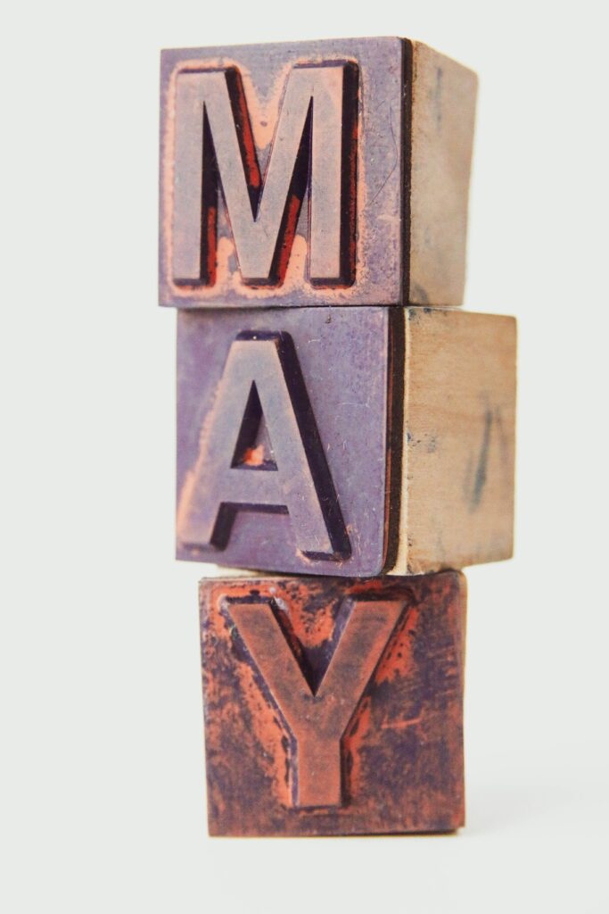 The image shows three wooden letterpress blocks stacked vertically, spelling the word "MAY." Each block has a letter carved into it, with a rustic and worn appearance. The background is plain and light-colored.