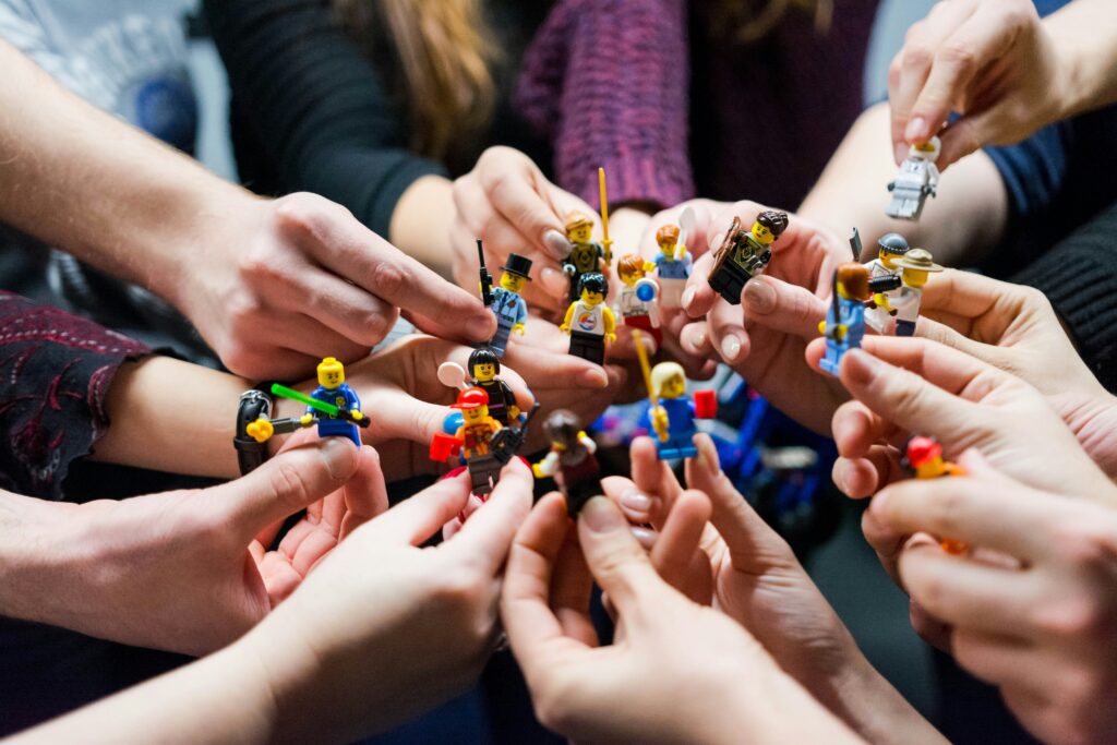 A group of people are holding various LEGO minifigures in their hands, forming a circle. The minifigures are diverse, featuring different outfits and accessories, and the hands are reaching in from all sides of the image.