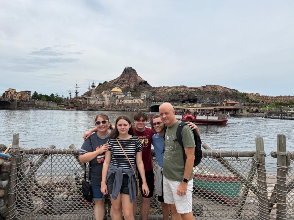 A group of five people is standing in front of a body of water with a scenic backdrop. Behind them, there is a rocky mountain with a castle-like structure and a ship. The people are smiling and posing for the photo, with some of them having their arms around each other. The scene appears to be at a theme park or tourist attraction.