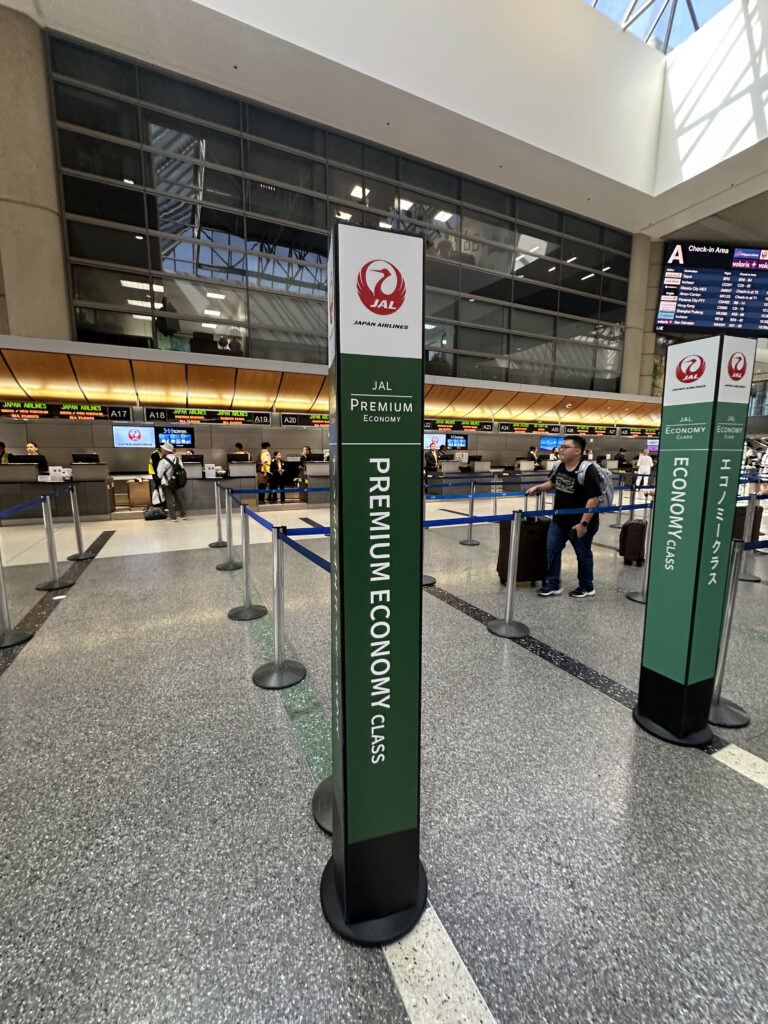 The image shows a Japan Airlines (JAL) check-in area at an airport. There are signs for "Premium Economy Class" and "Economy Class" with the JAL logo. The area is organized with stanchions and ropes to guide passengers. In the background, there are check-in counters with staff and digital screens displaying flight information. The ceiling is high with large windows allowing natural light to enter.