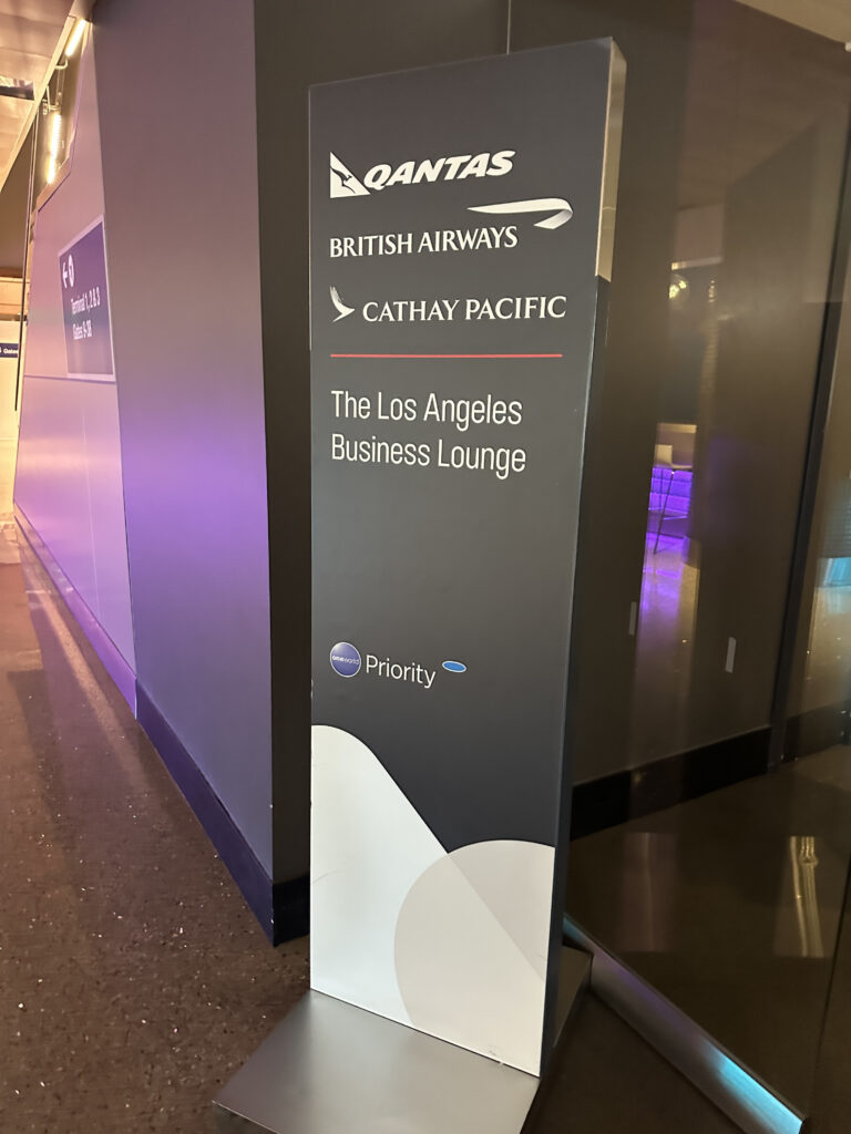 The image shows a sign for "The Los Angeles Business Lounge" featuring logos of Qantas, British Airways, and Cathay Pacific. The sign also includes the oneworld alliance logo and the word "Priority." The sign is located in an airport terminal, as indicated by the nearby directional sign for gates.