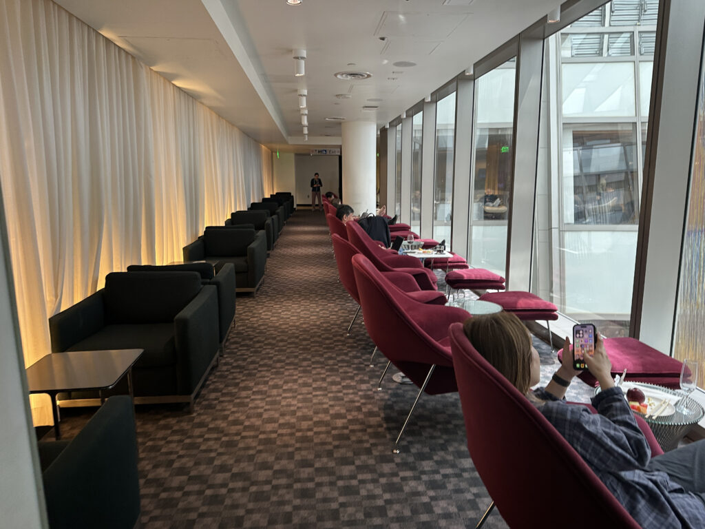 A modern lounge area with a row of black armchairs on the left and red lounge chairs with footrests on the right. The right side features large windows allowing natural light to enter. A person is seated in the foreground on a red chair, holding a smartphone. There are small tables with drinks and snacks next to the chairs. The space has a relaxed atmosphere with soft lighting from the left.