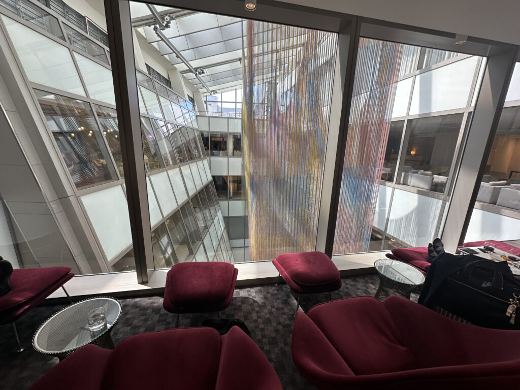 The image shows a modern indoor lounge area with large glass windows overlooking an atrium. The windows reveal a view of the building's interior architecture, featuring multiple floors with glass panels. Inside the lounge, there are plush, burgundy chairs and footrests arranged around small, round tables. A glass of water is on one of the tables, and a bag with a jacket is placed on a chair. The space is well-lit, with natural light streaming in through the windows.