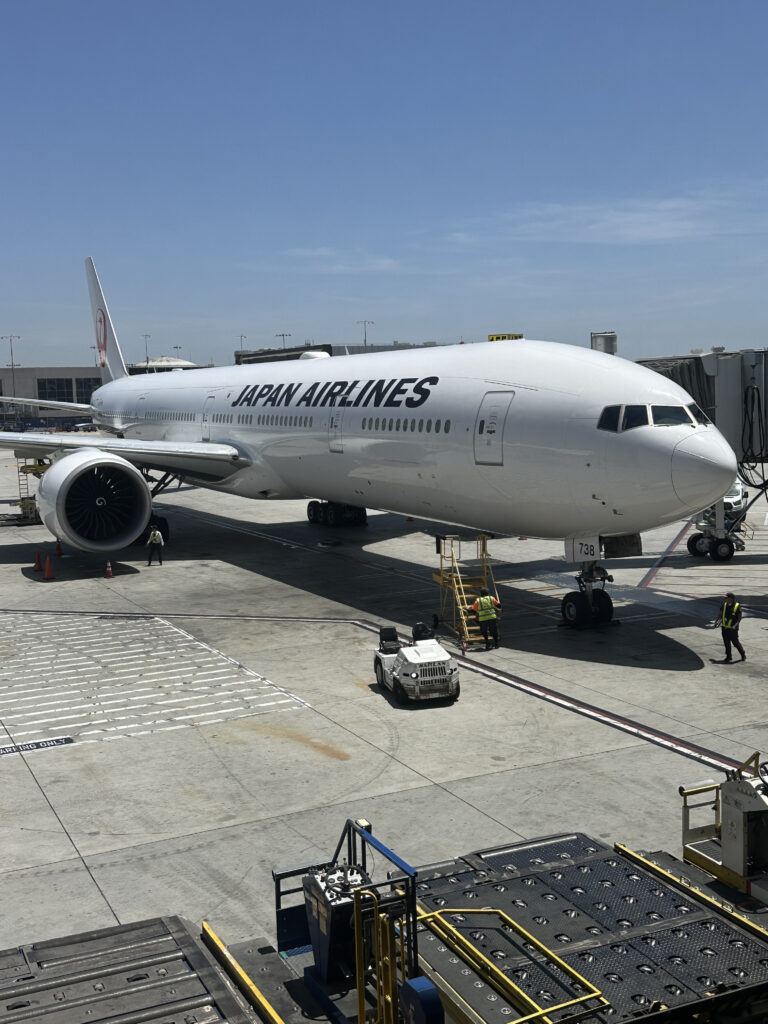 A Japan Airlines airplane is parked at an airport gate. The aircraft is white with "JAPAN AIRLINES" written on the side. Ground crew members and equipment are visible around the plane, and the sky is clear and blue.