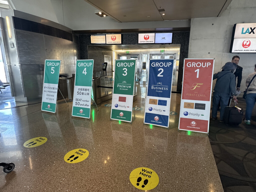 The image shows a boarding area at an airport with five signs indicating different boarding groups for Japan Airlines. The signs are labeled "Group 5" through "Group 1," with Group 1 being for JAL First Class and Group 2 for JAL Business Class. Group 3 is for JAL Premium Economy, Group 4 for seat numbers 50 and after, and Group 5 for all passengers. There are yellow floor markers with footprints and the text "Wait Here." People with luggage are visible in the background.