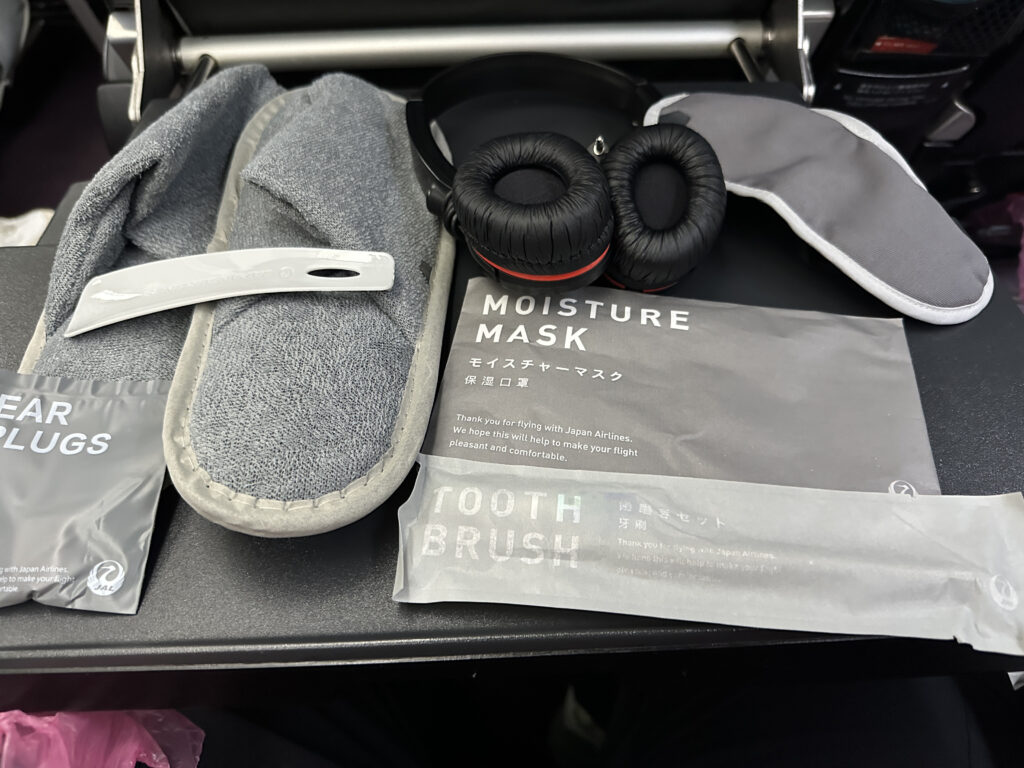 The image shows a collection of in-flight amenities on an airplane tray table. Items include a pair of gray slippers, a sleep mask, a pair of headphones, a moisture mask, ear plugs, and a toothbrush set. The packaging for the moisture mask and toothbrush set has text indicating they are provided by Japan Airlines.