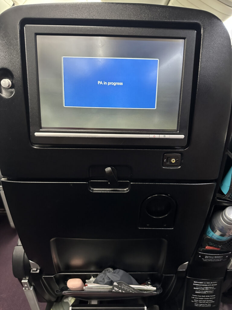 The image shows the back of an airplane seat with an in-flight entertainment screen displaying a blue message that reads "PA in progress." Below the screen, there is a tray table in the upright position, and a seat pocket containing various items, including a water bottle and some reading materials.