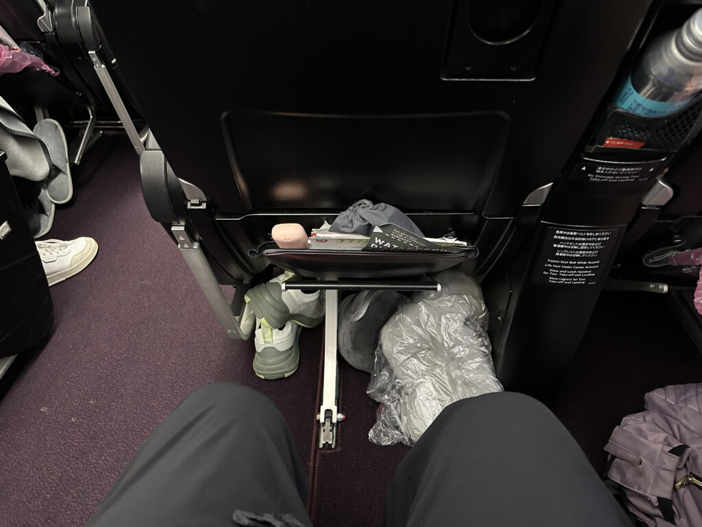The image shows the view from a passenger's seat on an airplane. The seatback in front has a pocket containing a magazine, a small pink object, and a gray item. Below, there are a pair of green and white sneakers, a plastic bag, and a gray item on the floor. The aisle is visible with a purple carpet, and there are other passengers' belongings, such as a purple bag, visible on the sides.