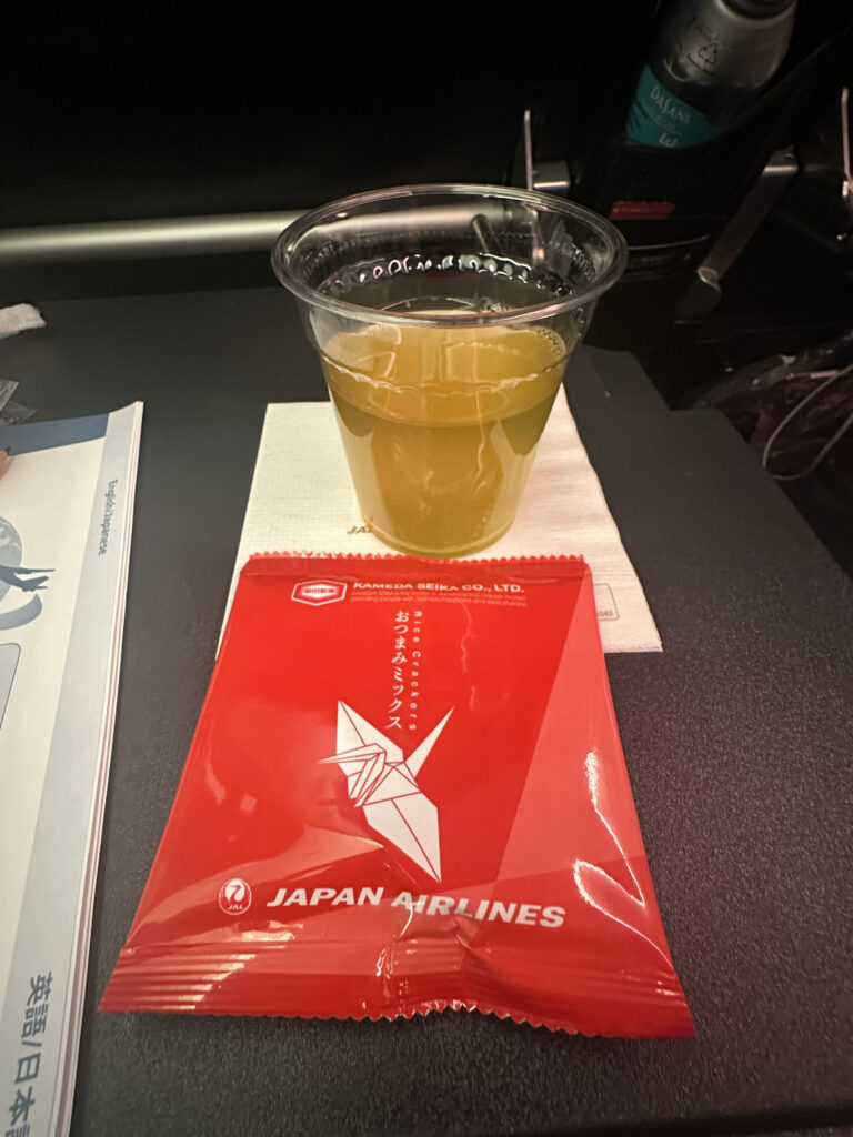 A plastic cup filled with a yellow beverage is placed on a white napkin on an airplane tray table. Next to it is a red packet labeled "Japan Airlines" with an origami crane design. A magazine or booklet is partially visible on the left, and a bottle is in the seat pocket in the background.