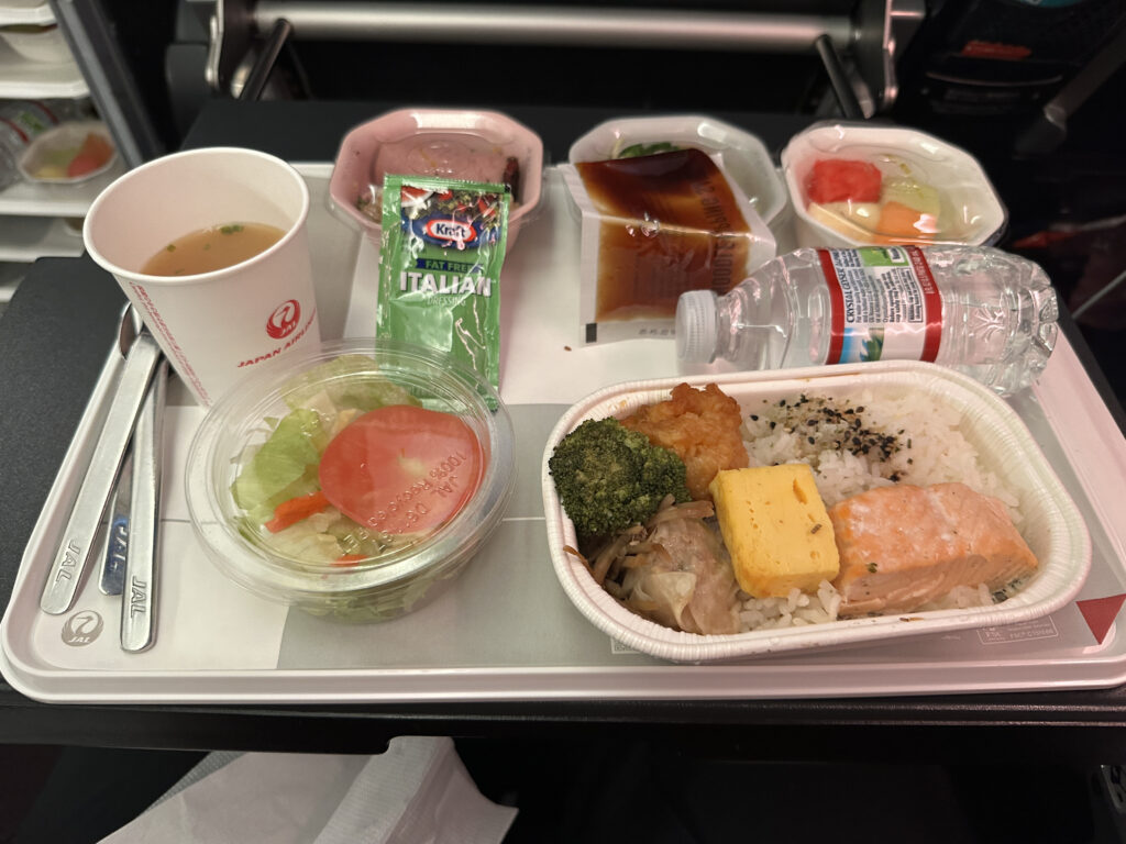 The image shows an airline meal on a tray. The meal includes a main dish with rice, a piece of salmon, a slice of tamagoyaki (Japanese omelette), broccoli, and a piece of fried food. There is a side salad with lettuce and a tomato slice, accompanied by a packet of Italian dressing. A cup of soup, a small bottle of water, and two dessert dishes with fruit and a sweet item are also on the tray. The tray and utensils are branded with the airline's logo.