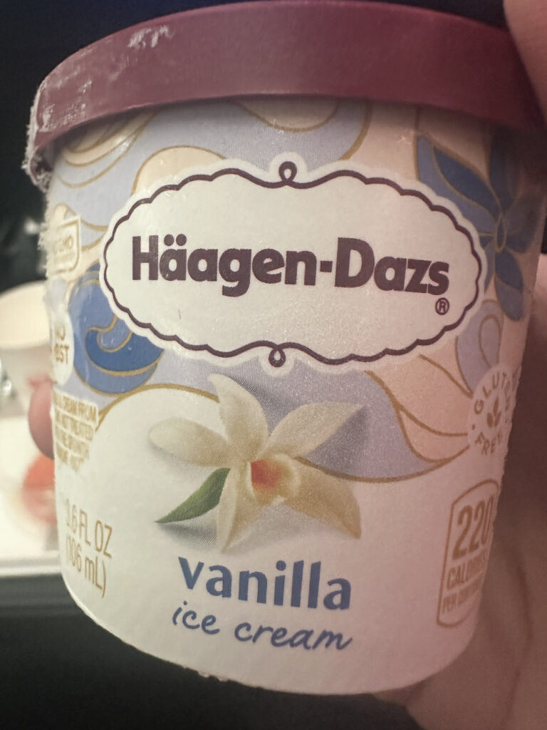 The image shows a small container of Häagen-Dazs vanilla ice cream. The container has a purple lid and features a design with a vanilla flower. The text on the container includes "Häagen-Dazs," "vanilla ice cream," and "220 calories per container." The container size is 3.6 fl oz (106 mL), and it is labeled as gluten-free.