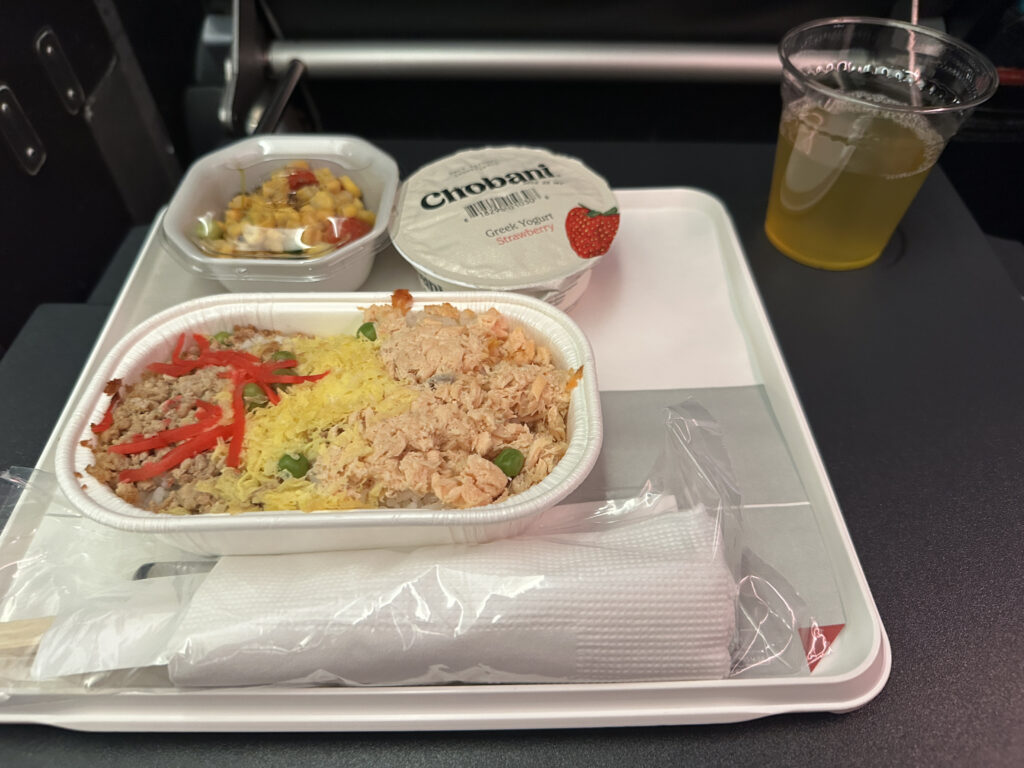 The image shows an airplane meal on a tray. The main dish is a container with rice topped with shredded meat, red peppers, and yellow garnish. There is a small container of mixed fruit or salad, a cup of Chobani Greek yogurt, and a plastic cup with a yellow beverage. A wrapped set of utensils is also on the tray.