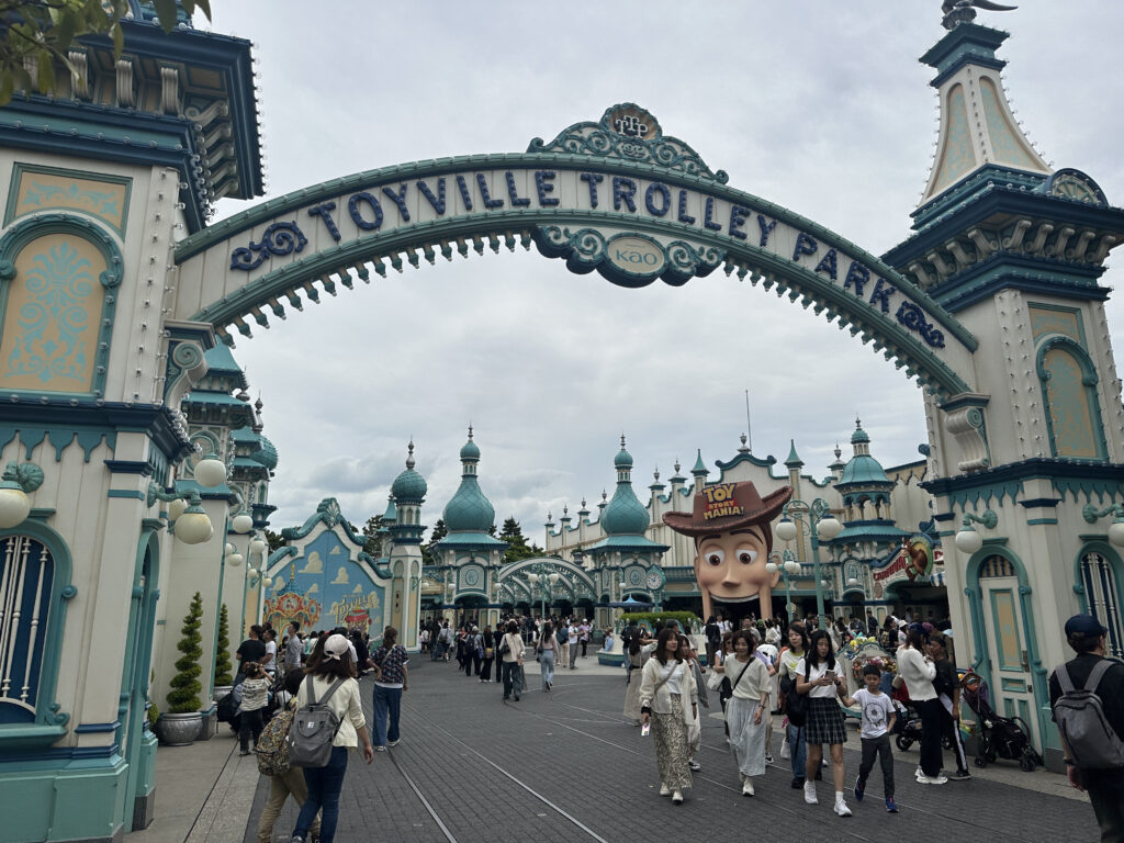 The image shows the entrance to "Toyville Trolley Park," a themed area with ornate, colorful architecture featuring domes and decorative elements. A large sign with the words "Toy Story Mania!" is visible, along with a giant figure of a character wearing a cowboy hat. Many people are walking around, enjoying the park.