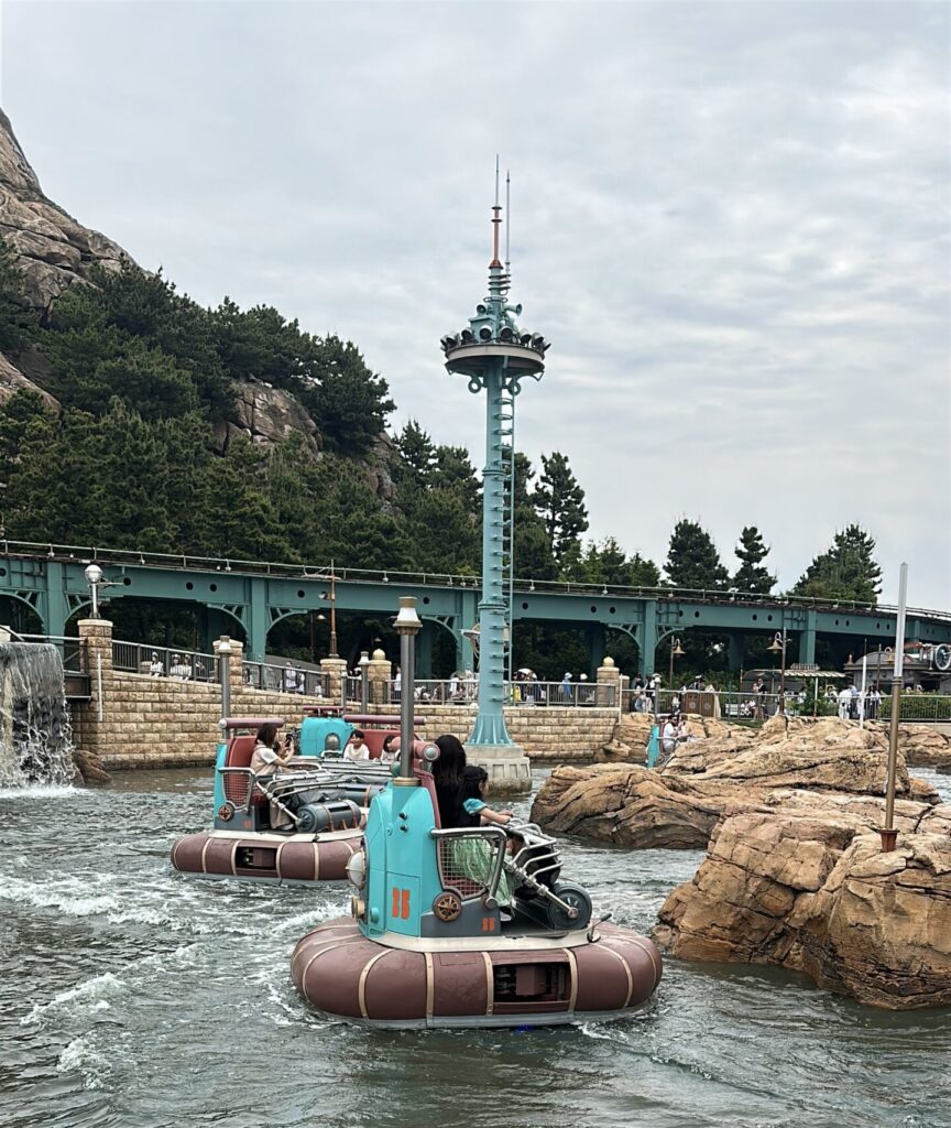 The image shows a water ride with several small, round boats carrying people. The boats are floating on a waterway surrounded by rocky terrain. In the background, there is a structure resembling a bridge and a tall, decorative pole. Trees and a rocky hill are visible in the distance under a cloudy sky.