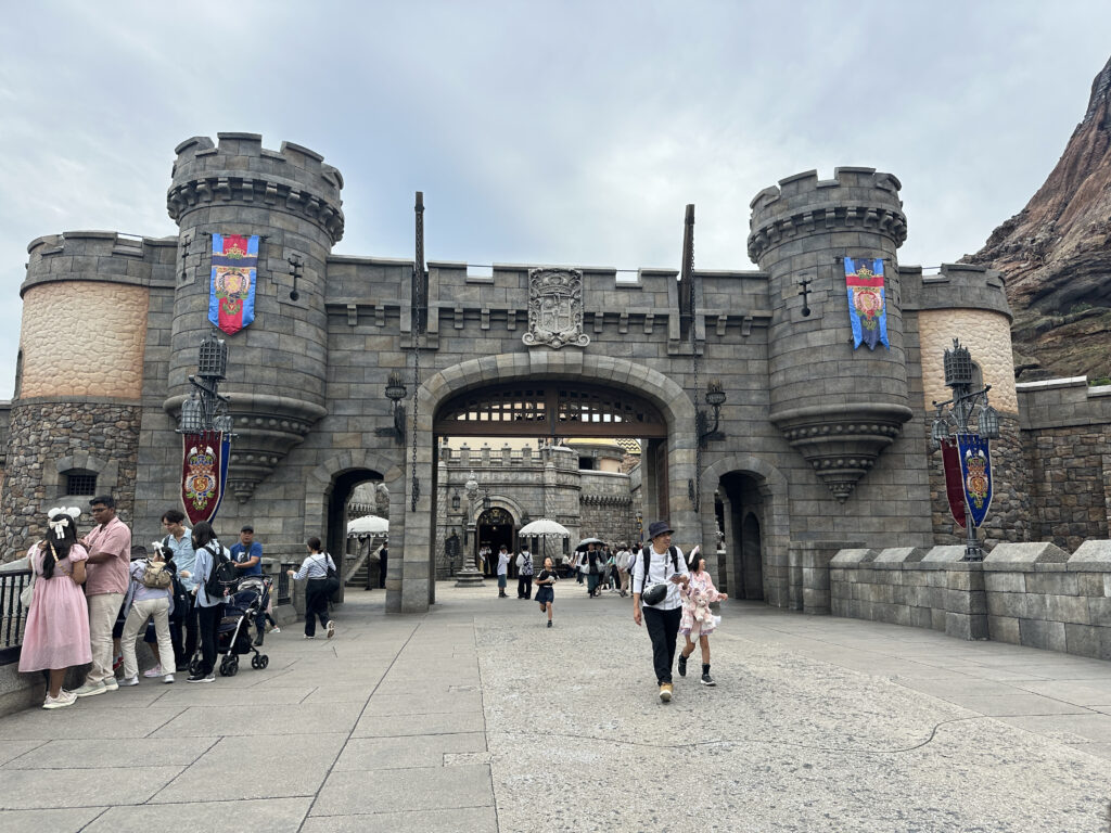 The image shows a stone castle entrance with two round towers on either side. The castle features decorative banners and a large archway leading inside. People are walking around the area, some with strollers and others taking photos. The sky is overcast, adding a dramatic backdrop to the scene.