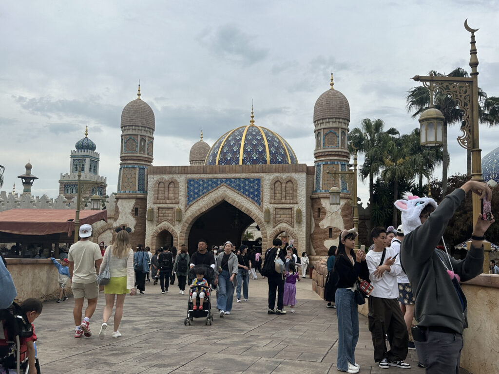 The image shows a lively scene at a theme park with a Middle Eastern architectural style. The building features ornate domes and towers with intricate blue and gold designs. People are walking around, some taking photos. One person is wearing a playful hat with animal ears. The sky is overcast, and there are palm trees in the background.