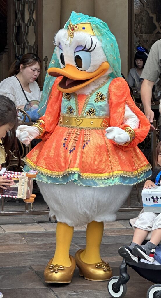 A character in a colorful costume is standing in an outdoor setting. The character is wearing an orange and turquoise outfit with gold embellishments and a matching headpiece. The character has large eyes and a beak, resembling a duck. People are around, including a child in a stroller and a person holding a toy.