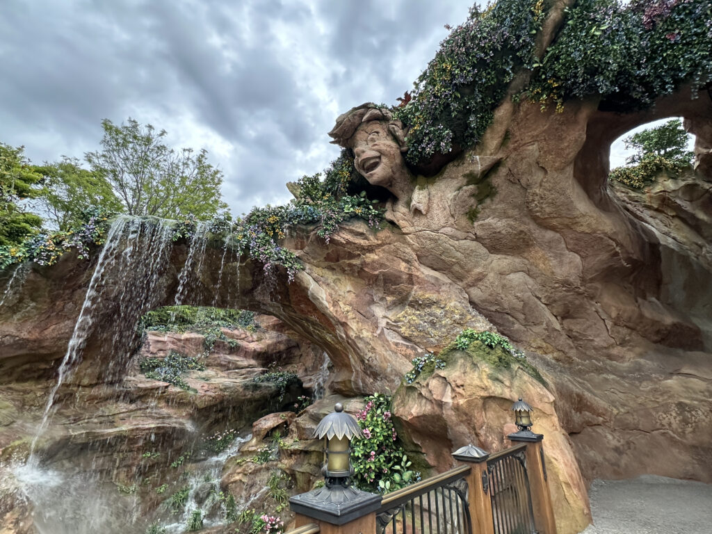 The image shows a rocky structure with a carved face of a smiling man integrated into the rock. Water cascades down from the top of the rock, creating a waterfall effect. The rock is adorned with greenery and flowers, adding a natural touch. In the foreground, there is a decorative fence with lanterns. The sky is overcast with clouds.