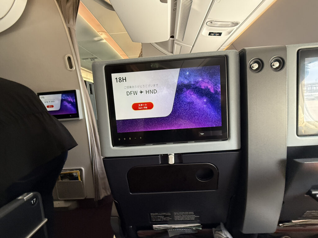 The image shows the interior of an airplane cabin, focusing on the back of a passenger seat equipped with an in-flight entertainment screen. The screen displays flight information, including the seat number "18H" and the route "DFW → HND." The background of the screen features a purple and black galaxy-like design. Another screen is visible on the seat to the left, showing similar content. The cabin appears to be in a premium or business class section, with spacious seating and a modern design.