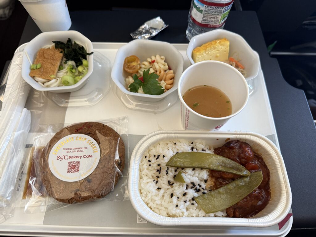 The image shows an airplane meal on a tray. The meal includes a main dish with rice, black sesame seeds, and a meat dish with sauce and snap peas. There are side dishes in small bowls, including noodles with green onions and seaweed, a pasta salad with a cherry tomato, and a piece of egg or cornbread. A cup of soup and a packaged chocolate chip cookie are also on the tray. A bottle of water and a cup are visible in the background.