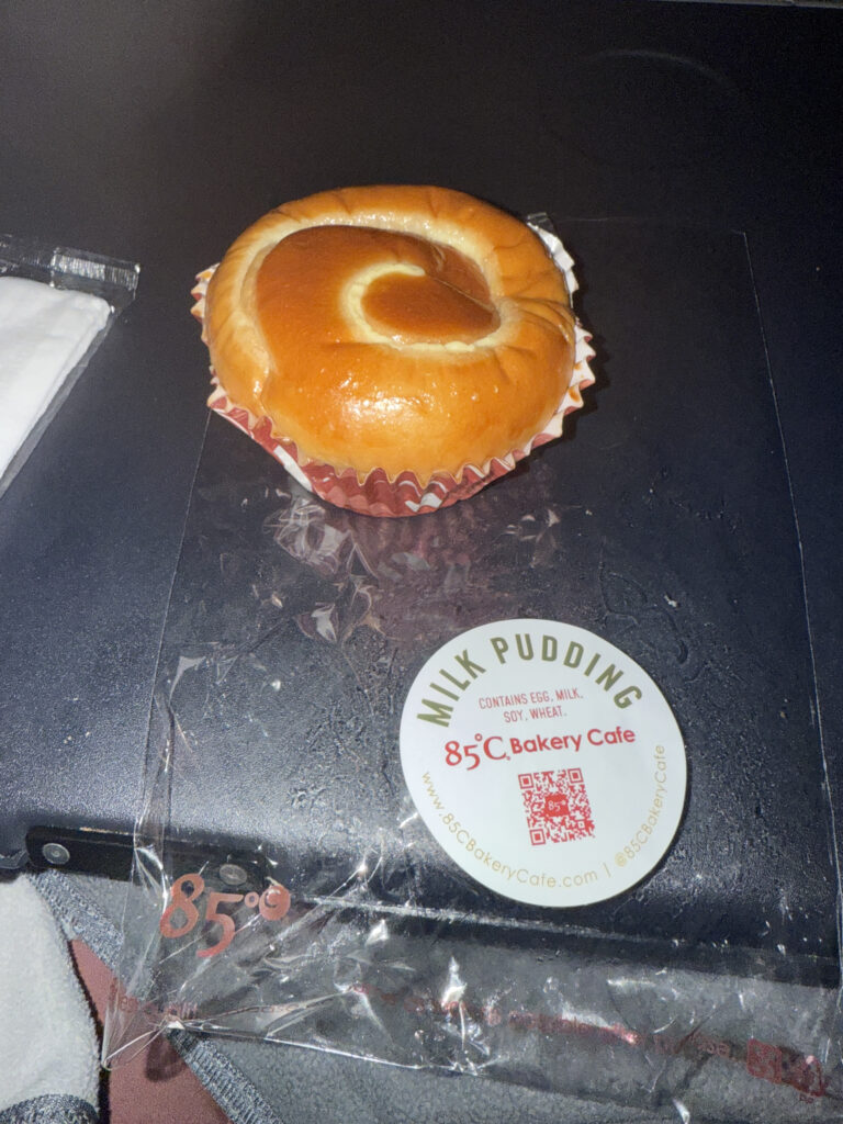 The image shows a milk pudding bun from 85°C Bakery Cafe, placed on a dark surface. The bun is golden-brown with a glossy finish and is sitting in a red and white paper cup. Next to it is a clear plastic packaging with a round label that reads "Milk Pudding" and includes the bakery's name and website.