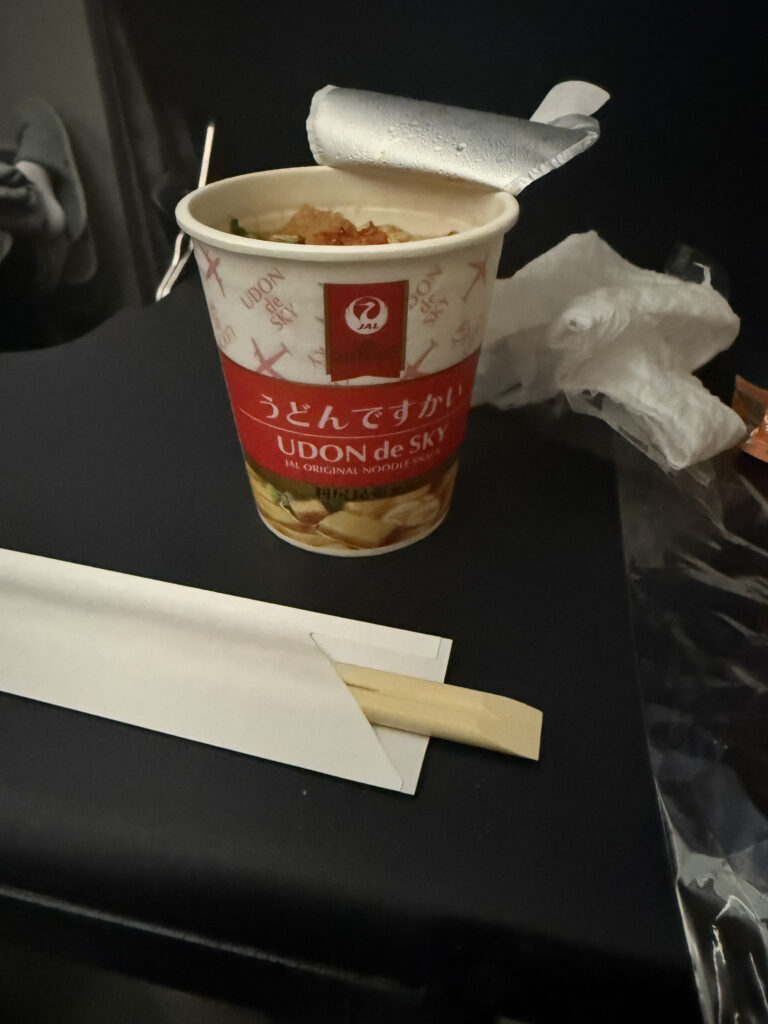 The image shows a cup of instant noodles labeled "UDON de SKY" on an airplane tray table. The foil lid is partially peeled back, revealing the noodles inside. A pair of wooden chopsticks is placed in a paper sleeve next to the cup. There is a crumpled napkin or tissue in the background.