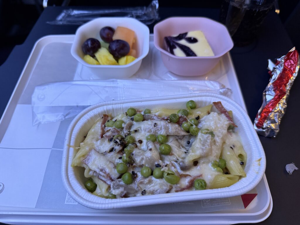 The image shows an airline meal on a tray. The main dish is pasta with peas, pieces of meat, and a creamy sauce. There are two side dishes: one with a mix of fruits including grapes, melon, and pineapple, and another with a dessert that appears to be a custard with a dark sauce on top. A drink is partially visible in the background, and there is a wrapped item, possibly a bread roll, on the side.
