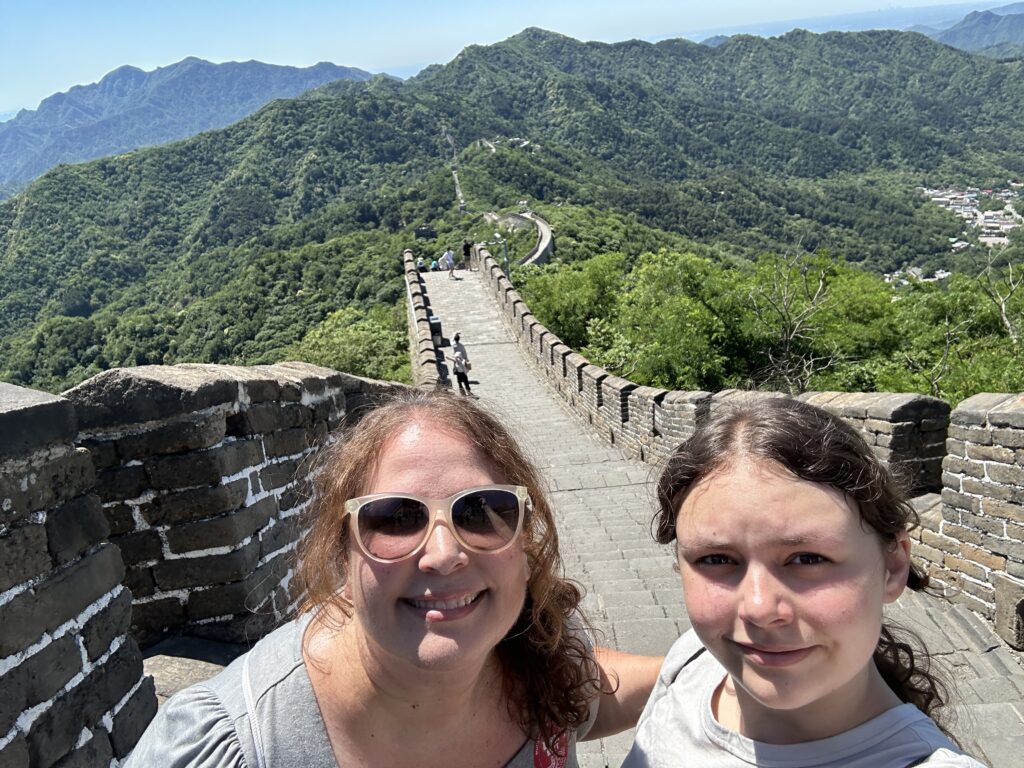 A selfie of two people on the Great Wall of China. They are smiling, with one wearing sunglasses. The wall stretches into the distance, surrounded by lush green mountains under a clear blue sky.
