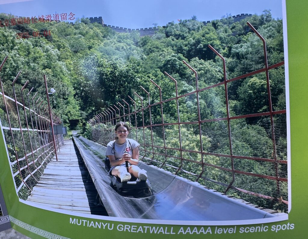 A person is riding a toboggan down a slide at the Mutianyu section of the Great Wall of China. The slide is surrounded by a metal fence, and lush green trees are visible in the background. The Great Wall can be seen at the top of the hill. The text at the bottom reads "MUTIANYU GREATWALL AAAAA level scenic spots."