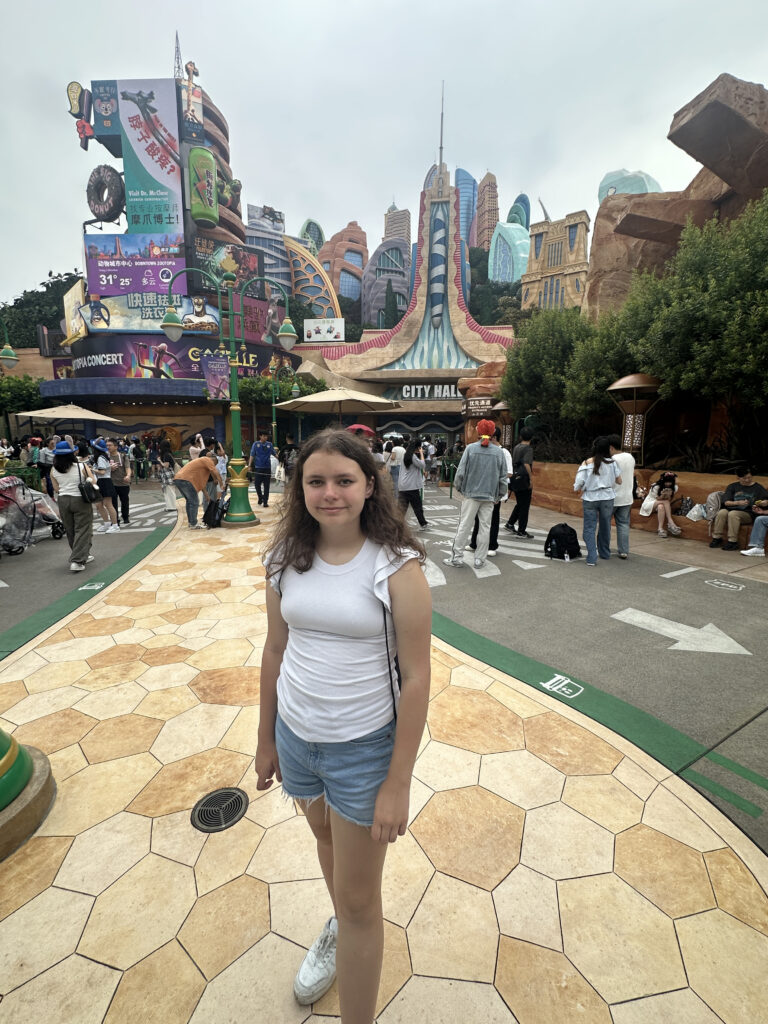 A young girl is standing on a patterned walkway in a theme park. She is wearing a white shirt, denim shorts, and white sneakers. Behind her, there are colorful, cartoon-like buildings with signs, including one that says "City Hall." The area is bustling with people walking around and taking photos. The sky is overcast.