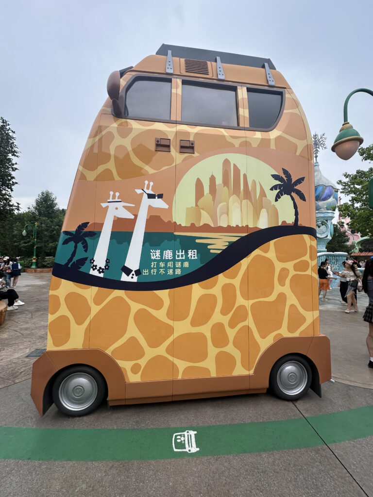 A vehicle designed to look like a giraffe, with a tall, boxy shape and giraffe-patterned exterior. The side features an illustration of two stylized giraffes wearing sunglasses, with a cityscape and palm trees in the background. There is text in a non-English language on the side. The vehicle is parked on a paved area with people and trees in the background.