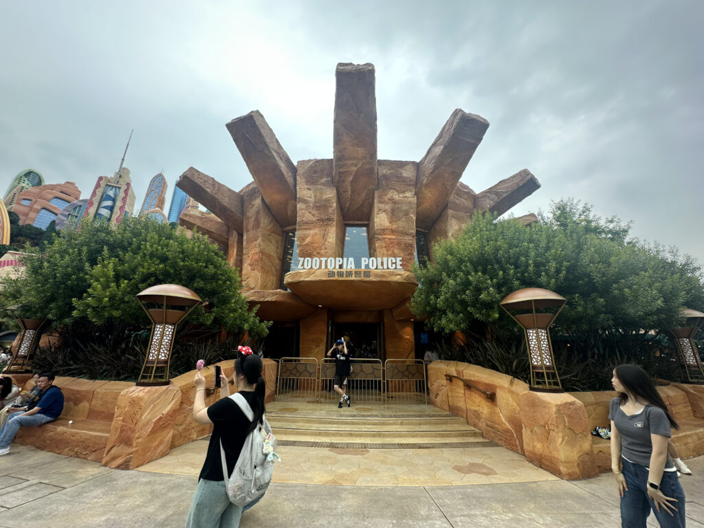 The image shows a building with a sign that reads "Zootopia Police" above the entrance. The structure has a rugged, stone-like appearance with large, protruding beams. There are trees and decorative lamps on either side of the entrance. Several people are in the foreground, with one person taking a photo and others walking or sitting nearby. The sky is overcast.