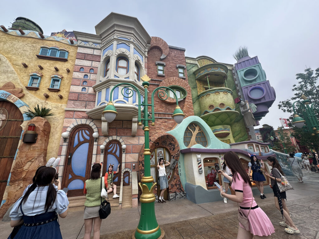 The image shows a whimsical, colorful street scene with a row of cartoonish, fantasy-style buildings. The architecture features a mix of shapes and colors, including yellow, pink, green, and purple facades with various decorative elements like round windows and ornate details. Several people are in the foreground, some taking photos and others posing. The atmosphere is lively and playful, suggesting a theme park or entertainment area.