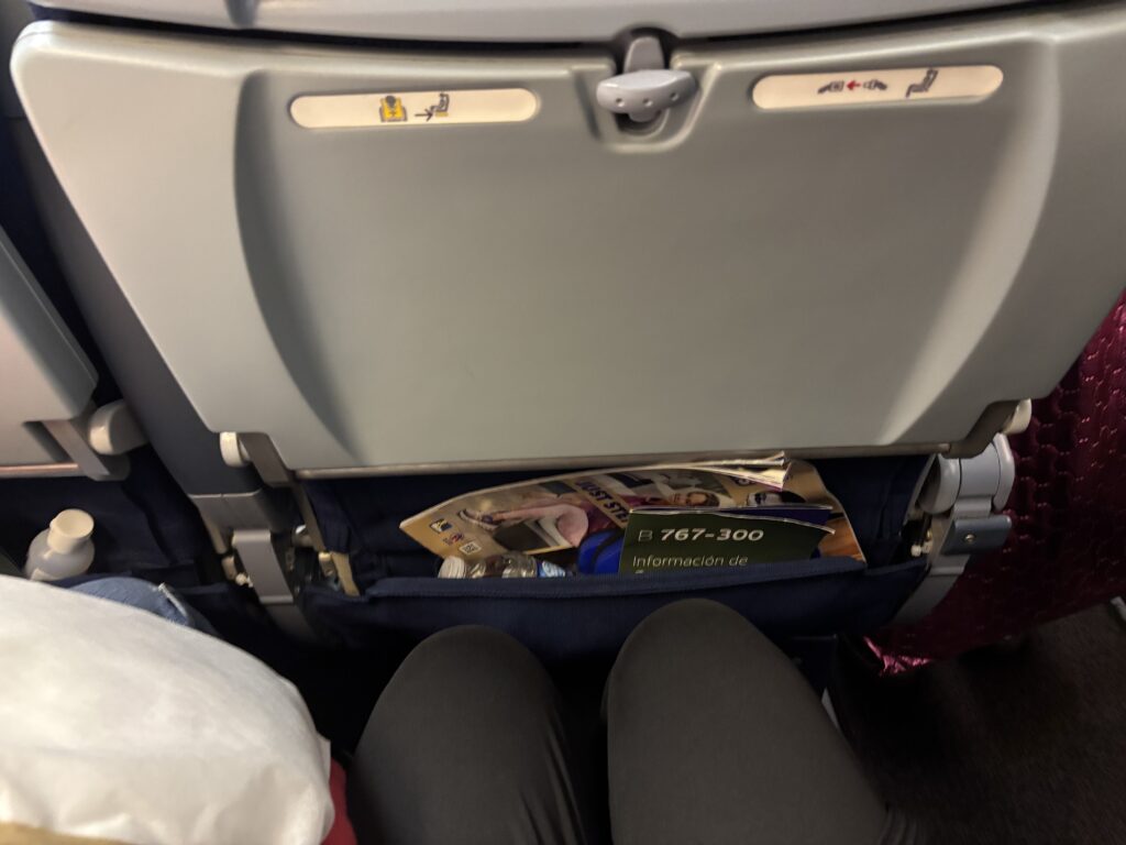 The image shows the back of an airplane seat with a tray table in the upright position. There are safety instructions and magazines in the seat pocket. A person's legs are visible in the foreground, and there is a bottle in the side pocket.