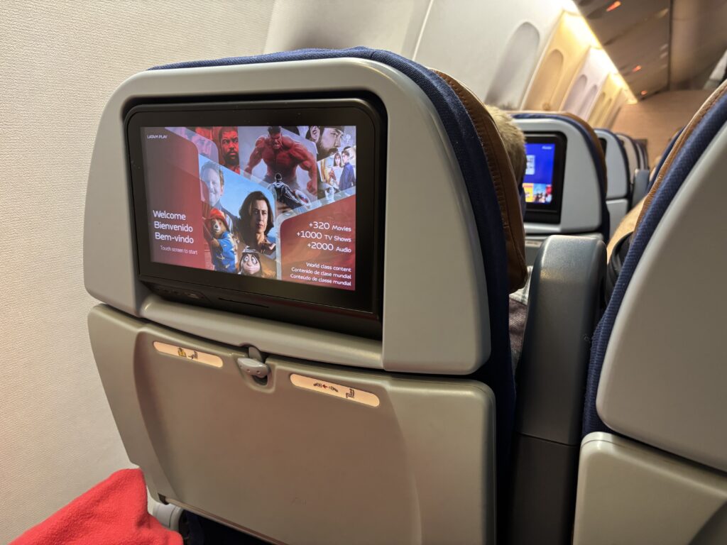 The image shows the back of an airplane seat with an in-flight entertainment screen. The screen displays a welcome message in multiple languages, including English, Spanish, and Portuguese, and offers options for movies, TV shows, and audio. The screen features images of various movie characters. The seatback tray table is in the upright position, and a red blanket is visible on the seat. The cabin interior and another screen on a different seat are visible in the background.